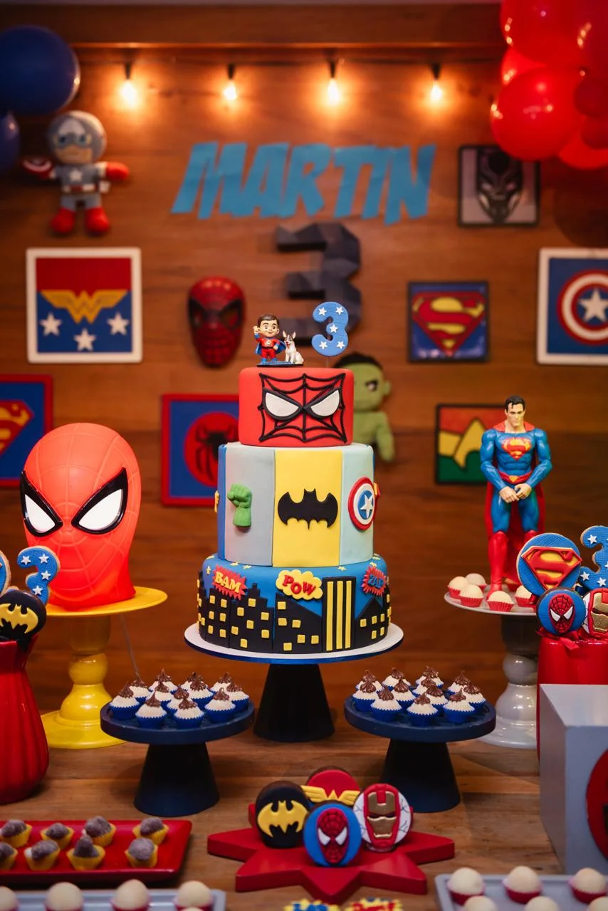 O bolo de aniversário visto de frente, com a miniatura do Martin no topo e a decoração de super-heróis da festa ao fundo