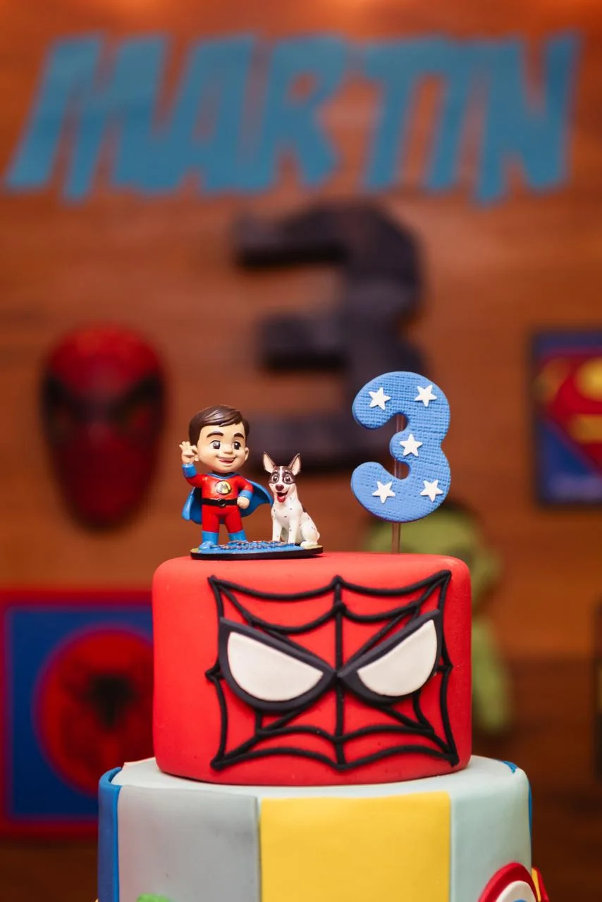 Close da miniatura personalizada do Martin como super-herói, de capa azul e traje vermelho, ao lado do cachorro da família, no topo do bolo de aniversário